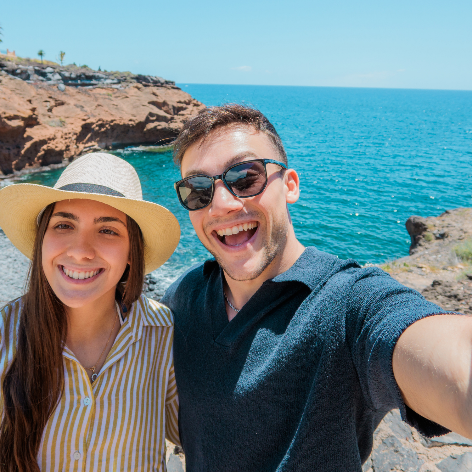 Two people smiling in front of the ocean in an image captured in a selfie style.